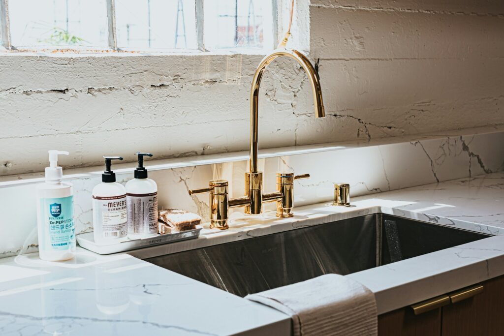 Modern kitchen sink with gold faucet, soap dispensers, and sponge on marble countertop, illustrating how to clean kitchen sink and drain effectively. Image by Andrew Valdivia via www.unsplash.com