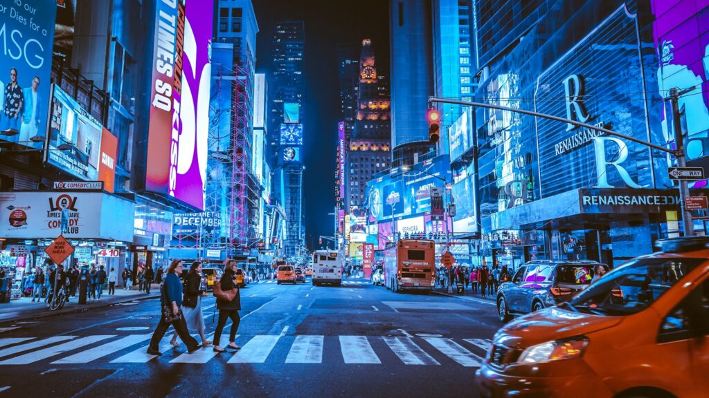 Times Square at night with bright billboards, traffic, and pedestrians crossing the street, reflecting the question: is living in New York expensive? Image by Andreas Niendorf via www.unsplash.com