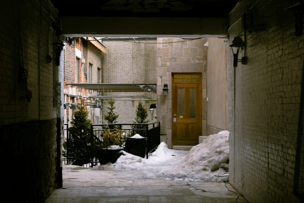 Snow piled beside a narrow urban walkway leading to a wooden door, highlighting the need for cleaning snow in winter conditions. Image by Jan Walter Luigi via www.unsplash.com